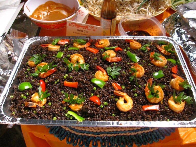 May include: A large rectangular foil tray of black rice, prawns, and vegetables. The prawns are cooked and arranged on top of the rice, with red and green peppers, peas, and parsley. Other dishes are visible in the background.