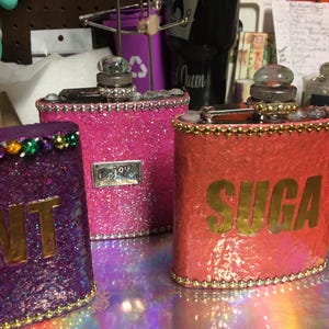 May include: Three glitter-covered flasks in pink, purple, and coral. The pink flask has a silver Enjoy label. The coral flask has the word SUGA in gold lettering. Each flask has a decorative top and is trimmed with rhinestones.