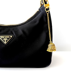 May include: Black Prada handbag with a gold zipper and logo. A gold chain with a decorative bell charm hangs from the top. The charm is gold with intricate detailing and small dangling beads.