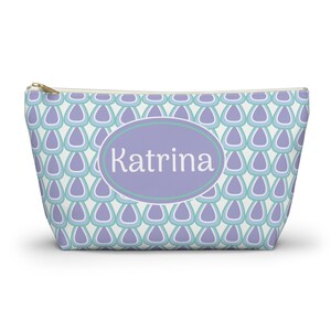 May include: A cosmetic bag featuring a pattern of blue, teal, and purple teardrop shapes on a white background. It has a gold zip and a purple oval with the name "Katrina" in white.