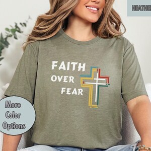 Puede incluir: Una camiseta de color verde oliva con el texto "FAITH OVER FEAR" y un gráfico de cruz colorido.