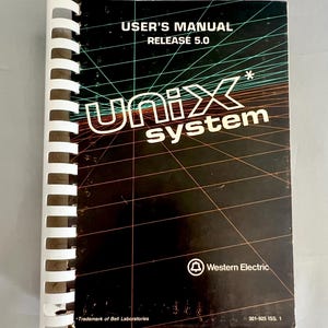 May include: A vintage "UNIX system" user's manual with a white spiral binding. The cover is black with the text "USER'S MANUAL RELEASE 5.0" and "UNIX system" in white. The Western Electric logo is visible.