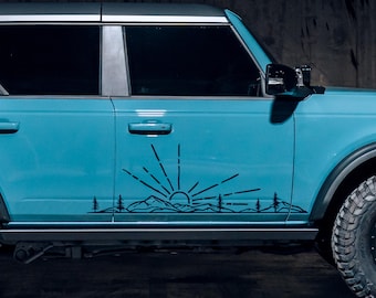 May include: A turquoise blue Ford Bronco with a black mountain range and sun decal on the side of the vehicle.