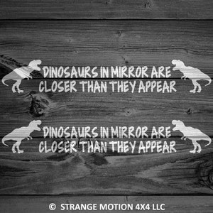 May include: Two white vinyl decals with a dinosaur silhouette and the text "Dinosaurs in mirror are closer than they appear" on a dark wood background.