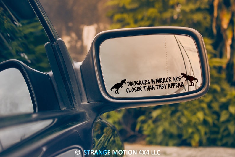 May include: A black car side mirror with a sticker that reads "Dinosaurs in mirror are closer than they appear" with a silhouette of a dinosaur on either side of the text.