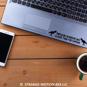May include: A laptop computer with a black and white sticker on the top that says "Dinosaurs in mirror are closer than they appear". The sticker features two dinosaur silhouettes. The laptop is on a wooden surface with a cell phone and a coffee mug.