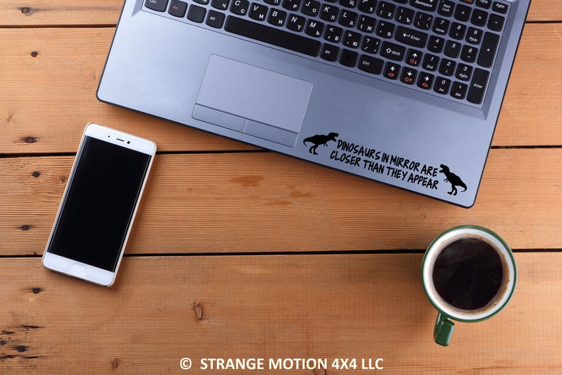 May include: A laptop computer with a black and white sticker on the top that says "Dinosaurs in mirror are closer than they appear". The sticker features two dinosaur silhouettes. The laptop is on a wooden surface with a cell phone and a coffee mug.