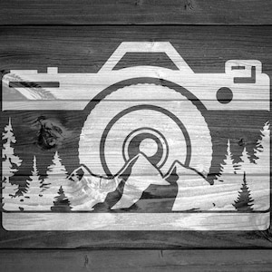 May include: A white silhouette of a camera with a lens that looks like a mountain range with trees on either side. The camera is on a woodgrain background.