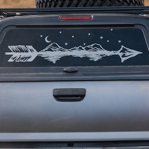 May include: A gray pickup truck with a rear window decal of a mountain range with a crescent moon and stars, and an arrow pointing through the mountains.