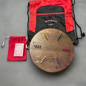 Könnte beinhalten: Ein rundes Holzspielbrett mit einem roten, blauen, grünen und weißen Steckspiel. Das Brett befindet sich in einem roten Kordelbeutel mit einer schwarzen Tasche, auf der "Sara Custom Products, really custom engraved products" steht. Das Spielbrett befindet sich auch in einem roten Kordelbeutel mit einem Kartenspiel.