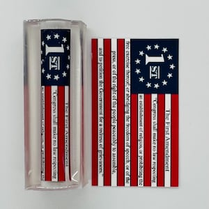 May include: A red, white, and blue American flag with the text "The First Amendment" and the text of the First Amendment. The flag is displayed on a white background.