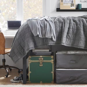 May include: A bedroom scene featuring a bed with a gray quilted comforter and a lighter gray blanket. Underneath the bed is a green trunk with gold accents and two gray storage bins. A desk with a laptop and a brown chair are visible.