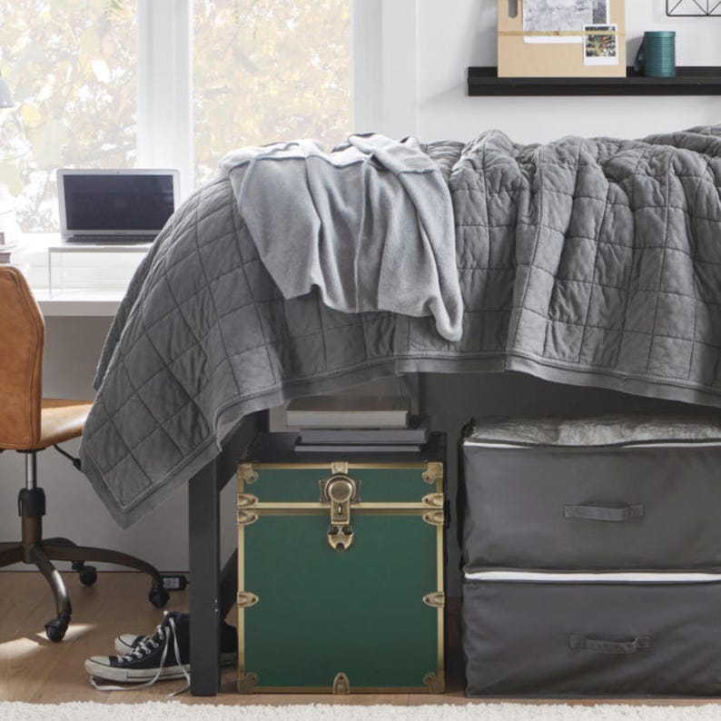 May include: A bedroom scene featuring a bed with a gray quilted comforter and a lighter gray blanket. Underneath the bed is a green trunk with gold accents and two gray storage bins. A desk with a laptop and a brown chair are visible.