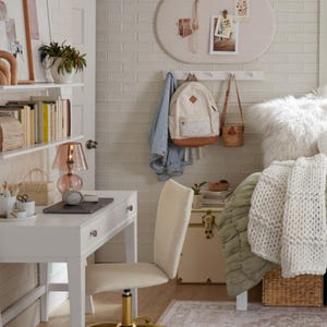 May include: A cozy bedroom scene with a white desk, chair, and shelves. A pink glass lamp sits on the desk next to a laptop. A backpack and purse hang on the wall. A bed with a white fluffy comforter and a woven basket are also visible.
