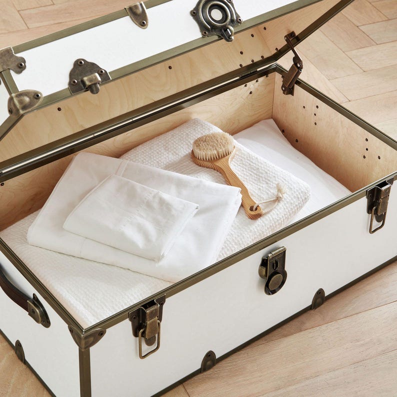 May include: A white trunk with brass-colored accents and hardware, open to reveal folded white linens and a wooden brush. The trunk's interior is lined with light-colored wood, and the trunk is sitting on a wooden floor.