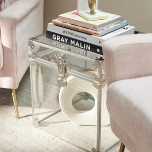 May include: A clear acrylic side table with silver metal trim and a trunk-style clasp. The table holds a stack of books, including "GRAY MALIN" and "AVEDON advertising." A white, circular decorative object sits inside the table.