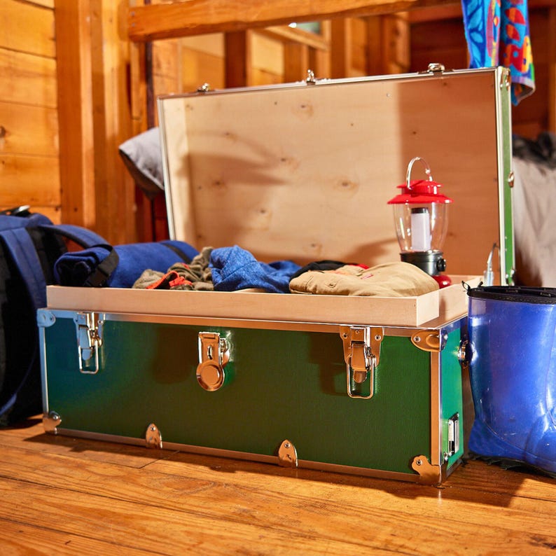 May include: A green trunk with silver hardware, open to reveal a wooden interior and contents including clothing and a red and black lantern. A blue backpack and blue boots are visible.