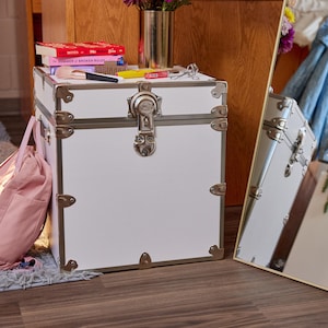 May include: A white storage trunk with silver hardware and corner protectors. The trunk has a latch closure and is topped with books, makeup, and a vase of flowers. A pink bag rests on the floor next to the trunk.