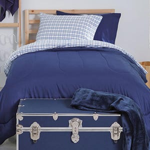May include: A navy blue bedding set with a plaid pillowcase and a matching comforter. A navy blue trunk with silver hardware sits at the foot of the bed, with a navy blue throw blanket draped over it. The bed frame is made of light-colored wood.