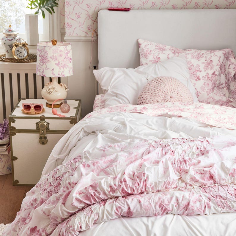 May include: A bedroom scene featuring a white bed with pink floral bedding, a white headboard, and a pink knitted pillow. A white trunk nightstand holds a lamp with a floral lampshade, sunglasses, and perfume. A gold alarm clock sits on a tray.