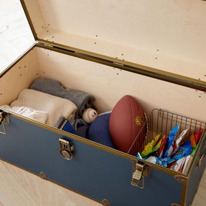 May include: A blue storage trunk with brass accents and a wooden interior. Inside are folded sweaters, a baseball, a football, and a wire basket filled with snacks. The trunk is open, revealing its contents.