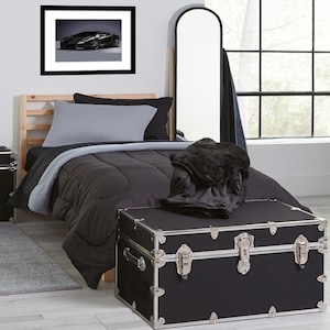 May include: A bedroom scene featuring a bed with gray and black bedding, a black trunk with silver accents, and a framed picture of a black sports car. A large mirror leans against the wall.