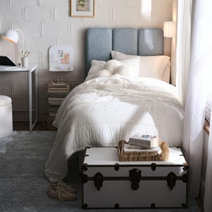 Puede incluir: Una escena de dormitorio con una cama con cabecero azul claro, ropa de cama blanca y un ba&uacute;l blanco al pie de la cama. Tambi&eacute;n se ven un escritorio con una l&aacute;mpara y una peque&ntilde;a pizarra. La habitaci&oacute;n tiene una est&eacute;tica acogedora y minimalista.