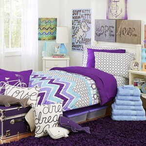 May include: A bedroom scene with a purple and blue color scheme. The bed has a patterned comforter and pillows, with a stack of blue towels. Decorative pillows and a trunk are in the foreground. Artwork and a lamp are also visible.