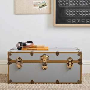 May include: A gray trunk with gold-colored hardware and accents. The trunk has a rectangular shape and is adorned with metal clasps and corner protectors. A pair of headphones and two books are on top of the trunk. The text "Vanishing World" is visible on one of the books.
