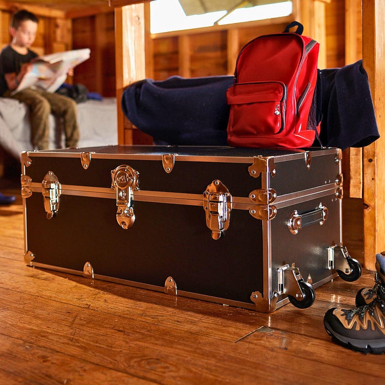 May include: A black trunk with gold-colored hardware and silver trim. The trunk has wheels and is rectangular. A red backpack and a navy blue blanket sit on top of the trunk. The trunk is on a wooden floor.