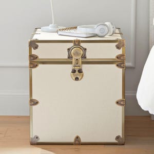 May include: Cream-colored trunk-style nightstand with brass-toned hardware. The nightstand features a rectangular shape with metal corner protectors and a central lock. A notebook and headphones rest on top.