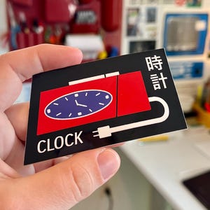 May include: A black and red sticker with a clock design. The sticker features a red rectangle with a blue clock face and the word "CLOCK" in white. Japanese characters are also present.