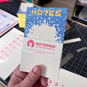 May include: A notepad with a blue pixelated design that reads "NOTES" in yellow. The notepad has 40 tear-off sheets and is printed and hand-assembled in Raleigh, NC. The notepad is held in a person's hand.