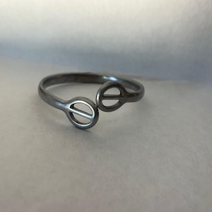 May include: A silver-toned metal bypass ring featuring two open circular terminals, each containing a horizontal bar across the center. The band has a smooth, polished finish and an open design that allows for adjustment to fit different finger sizes.