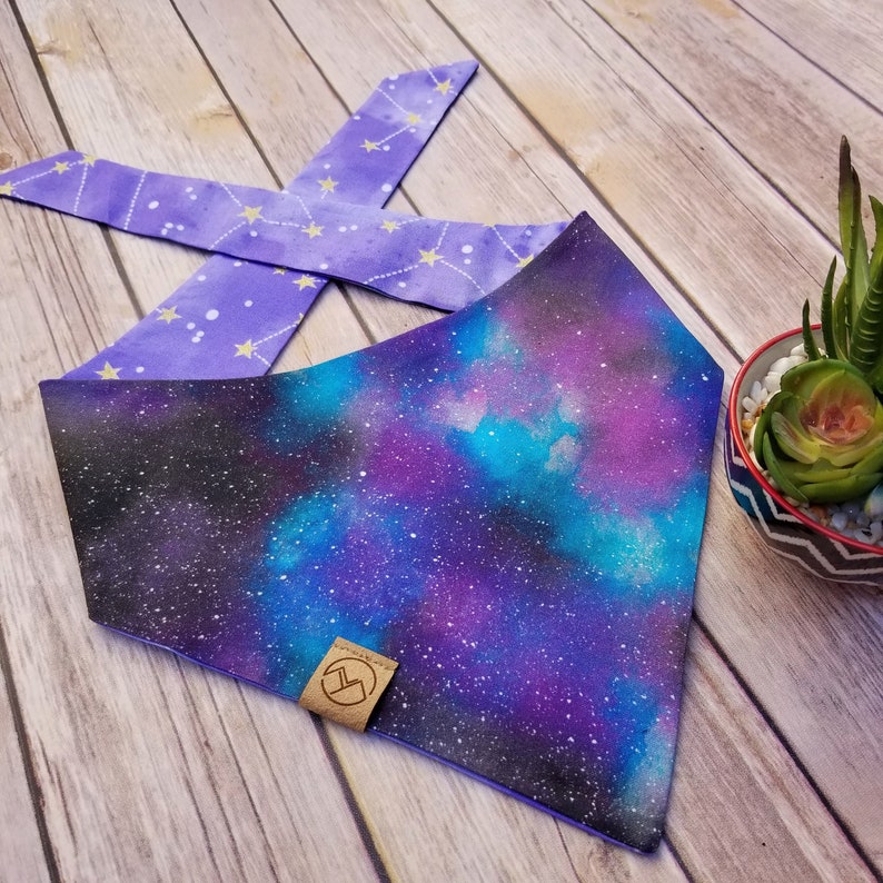 Puede incluir: Bandana para perro con estampado de galaxia p&uacute;rpura y azul con un estampado de estrellas doradas y p&uacute;rpuras en el reverso. La bandana tiene una etiqueta de cuero marr&oacute;n con un logotipo.