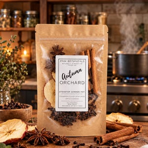 Op de afbeelding: Een bruine kraftpapieren zak met een stovetop simmer pot mix, gelabeld PNW Botanicals Autumn Orchard. Het doorzichtige venster toont gedroogde appelschijfjes, kaneelstokjes, steranijs en kruidnagels. Het etiket vermeldt de ingrediënten.