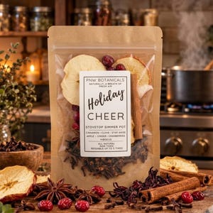 May include: A brown kraft paper pouch containing a stovetop simmer pot mix labeled PNW Botanicals Holiday Cheer. The clear window reveals dried apple slices, cinnamon sticks, star anise, cloves, cranberries, and hibiscus petals.
