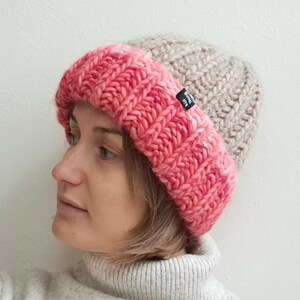 Könnte beinhalten: Eine gestrickte Mütze mit einem Farbverlauf von Pink zu Beige. Die Mütze hat eine dicke Strickstruktur und ein kleines schwarzes Etikett an der Seite.
