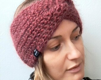 May include: A knitted headband in a dusty rose color. The headband is made of a thick, textured yarn and features a twisted knot detail at the top. A small black tag is sewn on the side. The person in the image is wearing a plaid shirt.
