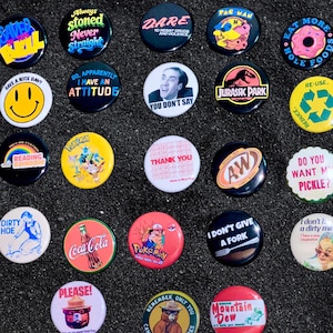 May include: A collection of twenty-one colorful pinback buttons with various designs and text. The buttons feature pop culture references, humorous phrases, and brand logos. The buttons are round and vary in size, with a black background.