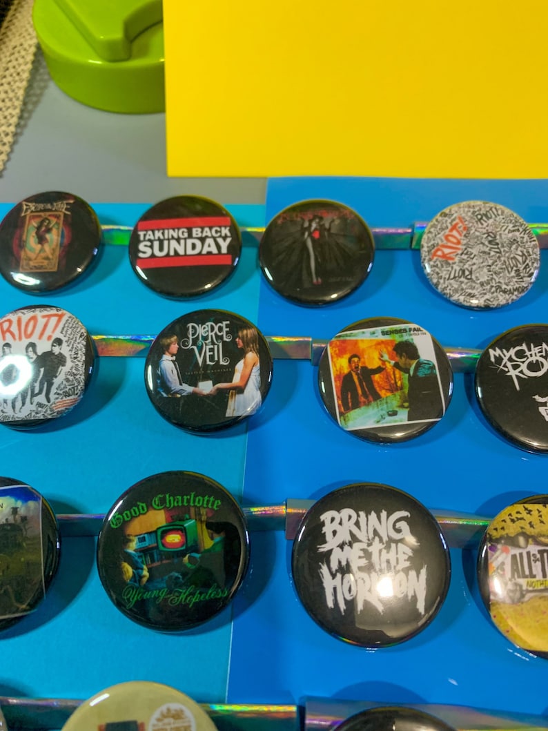 Puede incluir: Una colecci&oacute;n de 15 botones redondos en blanco y negro con logotipos de bandas. Los botones presentan bandas como Taking Back Sunday, Pierce the Veil, Bring Me the Horizon, Good Charlotte y Riot! Los botones se muestran sobre un fondo azul.