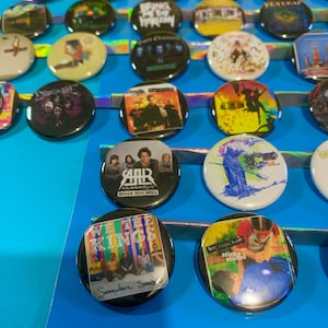 Puede incluir: Una colecci&oacute;n de botones redondos con varios logotipos de bandas y portadas de &aacute;lbumes. Los botones est&aacute;n dispuestos en filas sobre una superficie azul. Algunas de las bandas que aparecen son "We The Kings", "My Chemical Romance", "All Time Low" y "The Used".