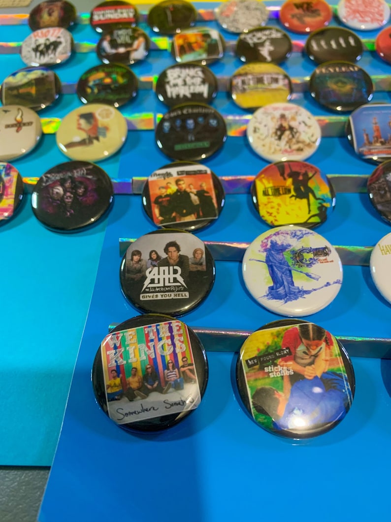 Puede incluir: Una colecci&oacute;n de botones redondos con varios logotipos de bandas y portadas de &aacute;lbumes. Los botones est&aacute;n dispuestos en filas sobre una superficie azul. Algunas de las bandas que aparecen son "We The Kings", "My Chemical Romance", "All Time Low" y "The Used".