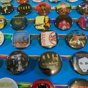 Puede incluir: Una colecci&oacute;n de botones redondos y coloridos con logotipos de bandas y portadas de &aacute;lbumes. Los botones est&aacute;n dispuestos en filas sobre una superficie azul. Algunas de las bandas que aparecen son The All-American Rejects, Sugarcult, Evanescence y Tonight Alive.