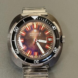 Può includere: Un orologio da polso automatico Junghans vintage con cinturino in acciaio inossidabile. Il quadrante dell'orologio presenta un design marrone e rosso con lancette e indicatori bianchi. La lunetta è nera con segni bianchi e la data è visualizzata alle 3.
