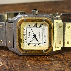 Puede incluir: Un reloj de cuarzo Geva vintage con una correa de metal plateado y dorado y una esfera cuadrada. La esfera del reloj es de color crema con manecillas negras y números romanos. El reloj se muestra sobre una superficie de madera.