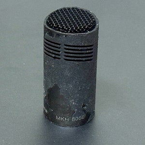May include: A black microphone with a metal grill and the text "MKH 8050" printed on the side.