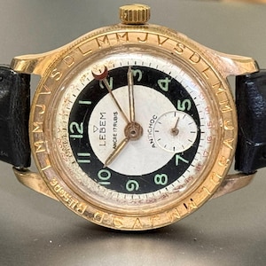May include: A vintage gold-tone wristwatch with a black leather strap featuring a crocodile texture. The round dial has a white center, a black outer ring with green Arabic numerals, and a gold bezel engraved with letters. Text reads LEBEM.