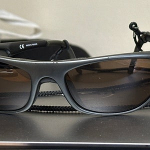 May include: A pair of black sunglasses with brown lenses and a black cord. The sunglasses have a CE mark and the words "Made in France" on the side.