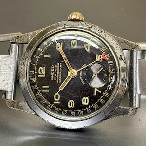 Może przedstawiać: Zegarek na rękę Anker w stylu vintage z czarną tarczą, złotymi wskazówkami i srebrną metalową bransoletą. Tarcza zegarka posiada cyfry arabskie i małą tarczę pomocniczą. Zegarek ma zużyty wygląd.